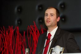 Ein Mann im Anzug hält eine Rede; im Hintergrund rote Dekoration.