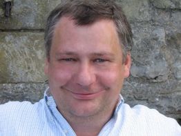 Eine lächelnde Person vor einer Steinmauer in einem hellen Hemd.