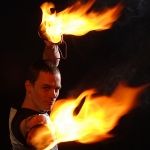 Mann mit zwei brennenden Fackeln, der sie kunstvoll bewegt, vor dunklem Hintergrund.