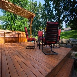 Wooden deck with red chairs and a shade structure, surrounded by trees and stones.