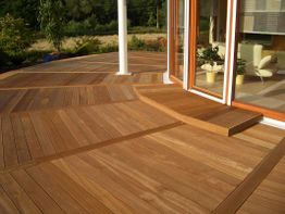 Holzterrasse mit Schrittstufe und großen Fenstern, umgeben von Pflanzen und Bäumen.