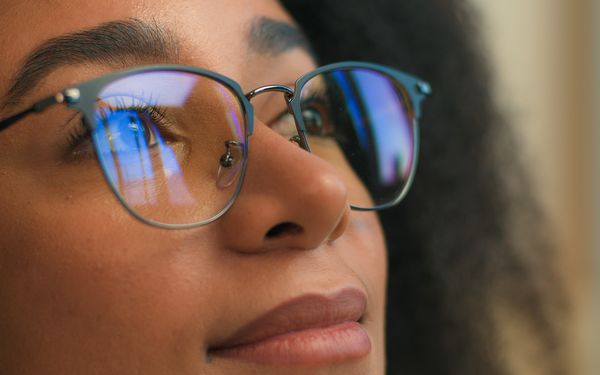Eine Frau hat eine Blaulichtfilterbrille auf und schaut auf einen Bildschirm der vor ihr steht. Das Licht des Bildschirms spiegelt sich in ihrer Brille.