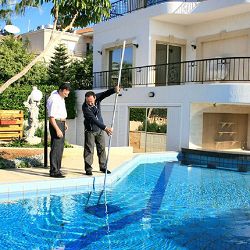 Two men cleaning a swimming pool with a net, in front of a white house and green garden.