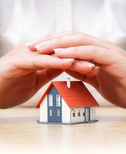 Manos protegen una casa en miniatura sobre una mesa de madera.