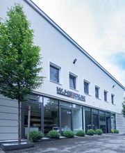 Modernes Bürogebäude mit großen Fenstern, davor ein Baum und Büsche, blauer Himmel.