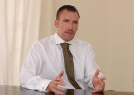 Mann in weißem Hemd und Krawatte sitzt am Tisch und gestikuliert vor einem beigen Hintergrund.