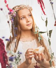 Niña con corona de flores, mirando al cielo, sosteniendo una concha entre flores colgantes.