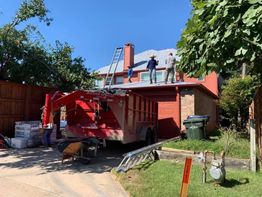 Workers repair a red house roof with a ladder; red trailer and tools nearby.