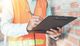 Person in safety vest writing on clipboard at construction site.
