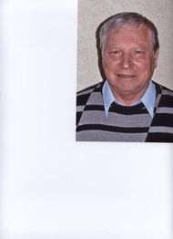 Älterer Mann lächelt in die Kamera, trägt gestreiften Pullover und Hemd, vor neutralem Hintergrund.