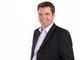 Ein lächelnder Mann im Anzug vor weißem Hintergrund.