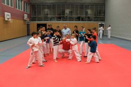 Kinder in Karate-Anzügen machen Kampfposen auf einer roten Matte in einer Turnhalle.