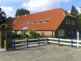 Rotes Backsteinhaus mit rotem Dach, blauer Zaun und Parkplatz im Vordergrund, bewölkter Himmel.