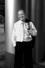 Mann in gestreiftem Hemd mit Jacke über der Schulter steht lächelnd vor einer Säule.