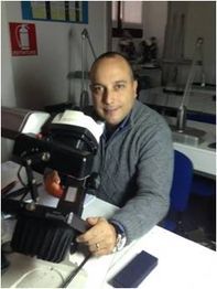 Uomo seduto al tavolo con uno strumento tecnico in mano, in ambiente di lavoro al chiuso.