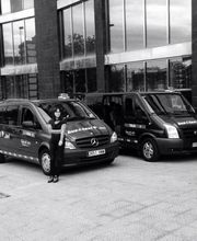 Dos taxis Mercedes Vito y Ford Transit estacionados frente a un edificio; una persona al lado izquierdo.