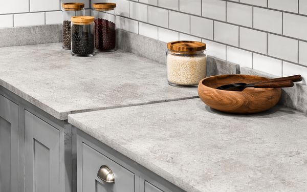 Encimera de cocina de hormigón en tono gris claro con acabado mate y textura ligeramente porosa, acompañada de tarros de vidrio con alimentos y un bol de madera sobre muebles blancos y salpicadero de azulejos.