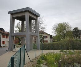 Grau-betonierter Pavillon neben einem Weg, umgeben von Häusern und Bäumen an einem bewölkten Tag.