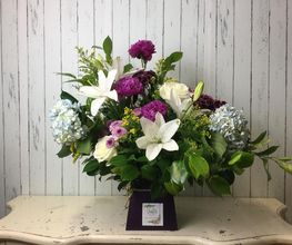 Ramo de flores con lirios blancos y flores moradas sobre una mesa. Fondo de madera clara.