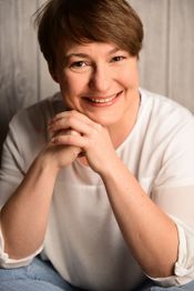 Eine lächelnde Person mit kurzem braunem Haar und weißem Hemd sitzt vor einer Holzwand.