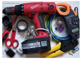 Trapano elettrico, forbici, pinze e multimetro su un tavolo con cavi e lampadine.
