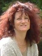 Eine Frau mit lockigem Haar lächelt draußen vor einem grünen Hintergrund.