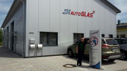 Gebäude mit JK Autoglas-Logo, Mann steht neben einem Schild vor einem geparkten Auto.