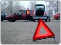 Warndreieck auf Straße, Auto mit geöffnetem Kofferraum, Person im Hintergrund bei einem Wagen.