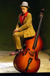 Eine Person in Clownkostüm sitzt auf einem großen Kontrabass, der auf einer Bühne steht.