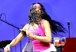 Frau tanzt energetisch in Pink vor blauem Hintergrund, Haare wehen im Schwung, Musik im Hintergrund.