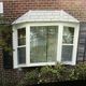 Bay window with white frame on a brick wall, adorned with dark shutters and leafy plants.
