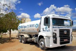 Caminhão-pipa branco de empresa de água estacionado em terreno árido sob céu azul com nuvens.