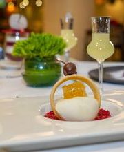 Postre gourmet con bebida amarilla en mesa elegante, adornado con planta y decoración de fondo.