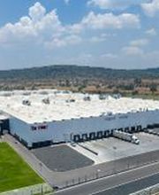 Vista aérea de una planta industrial blanca, rodeada de áreas verdes y un estacionamiento amplio.