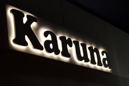 Letrero iluminado con la palabra "Karuna" en letras grandes y oscuras sobre fondo luminoso.