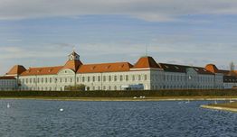Großes historisches Gebäude mit rotem Dach vor einem See unter blauem Himmel.