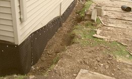 House foundation with black waterproofing membrane and nearby dug trench in progress.