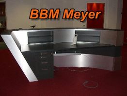 Halbrunder Empfangstresen mit Schrankelementen auf rotem Teppichboden. Text: BBM Meyer.
