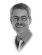 Schwarz-weiß Porträt eines lächelnden Mannes mit Brille und Krawatte vor weißem Hintergrund.