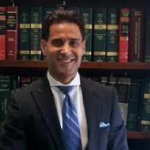 Man in a suit smiling in front of a bookshelf with legal books.