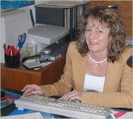 Frau mit lockigem Haar sitzt lächelnd an einem Schreibtisch mit Tastatur und Büromaterial.