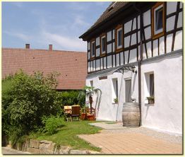 Fachwerkhaus mit Terrasse, Tischgruppe und Weinfass, umgeben von Garten und anderen Gebäuden.