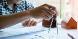 Hand using compass on architectural plans, with scale model houses in the background.