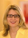 Frau mit Brille und blondem Haar, lächelt in die Kamera, trägt ein gelbes Hemd.