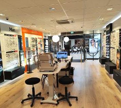 Optiekwinkel met brilrekken, een tafel en stoelen in een moderne en lichte ruimte.