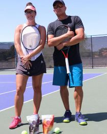 Two people on a tennis court holding rackets, standing near tennis balls and bags.