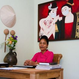 Frau in rosa Bluse sitzt lächelnd an einem Tisch, hinter ihr ein buntes Gemälde mit Masken.