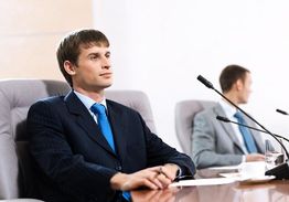 Man in suit at a conference table with microphones, another man in background.