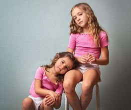 Dos niñas con blusas rosas, una sentada en un taburete y la otra apoyada en su rodilla.