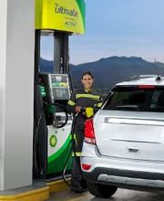Mujer en estación de servicio BP Ultimate cargando combustible a un coche blanco, montaña al fondo.